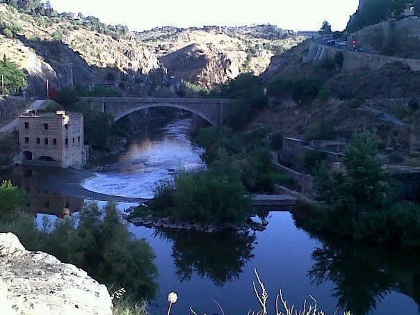 Puente de piedra sobre un río, con edificios en las orillas y terreno montañoso al fondo.