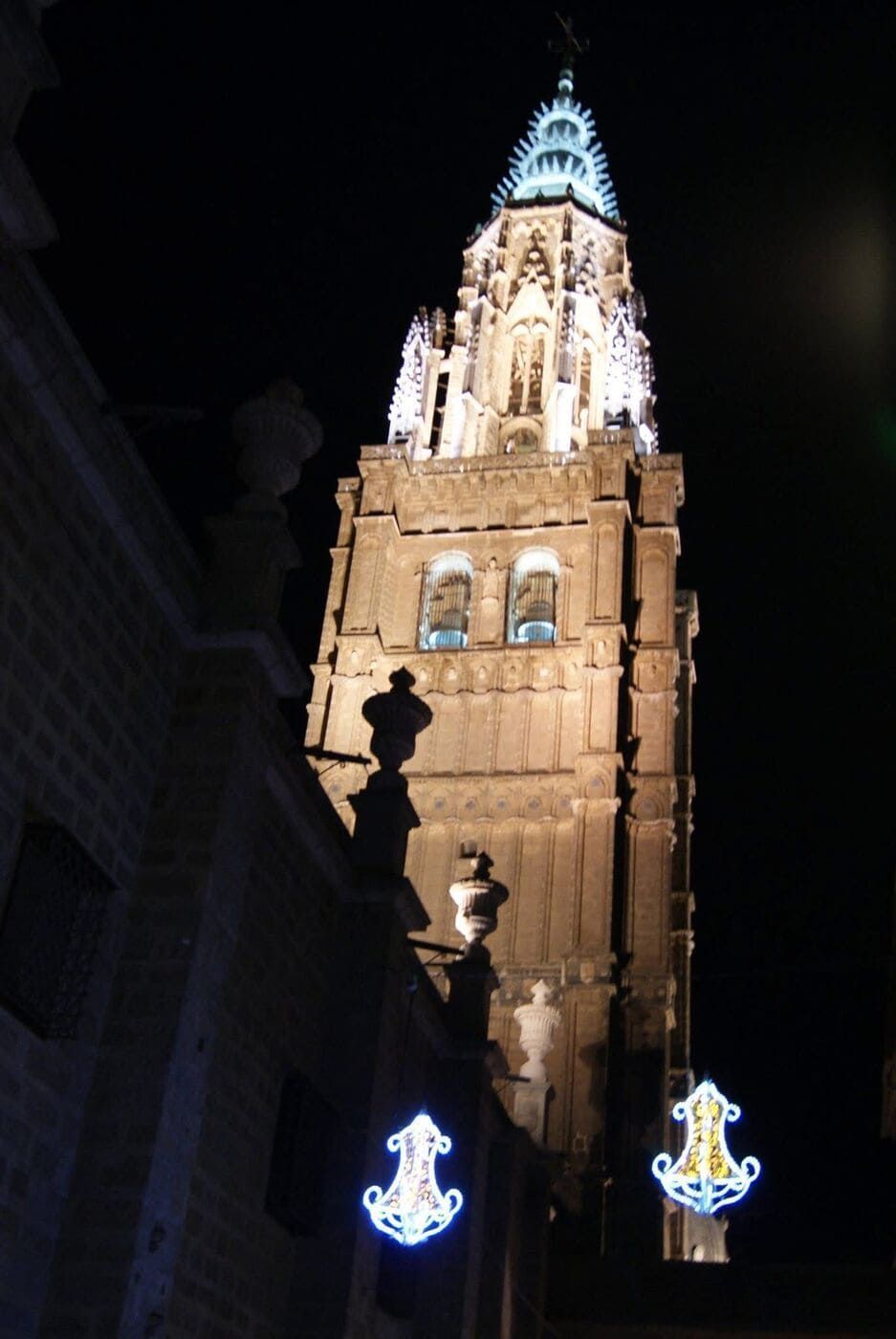 Vista nocturna de la torre de la catedral iluminada, con su ornamentada mampostería. Decoraciones de luz azul cuelgan debajo.
