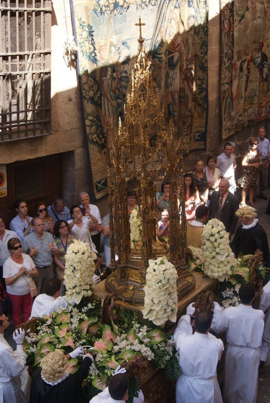 Procesión religiosa con custodia dorada ornamentada, portada por personas vestidas de blanco. La multitud observa, con un tapiz como telón de fondo.