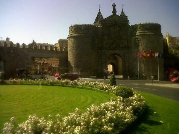 Puerta histórica de la ciudad con torres redondeadas y entrada arqueada, césped y arbustos en flor.