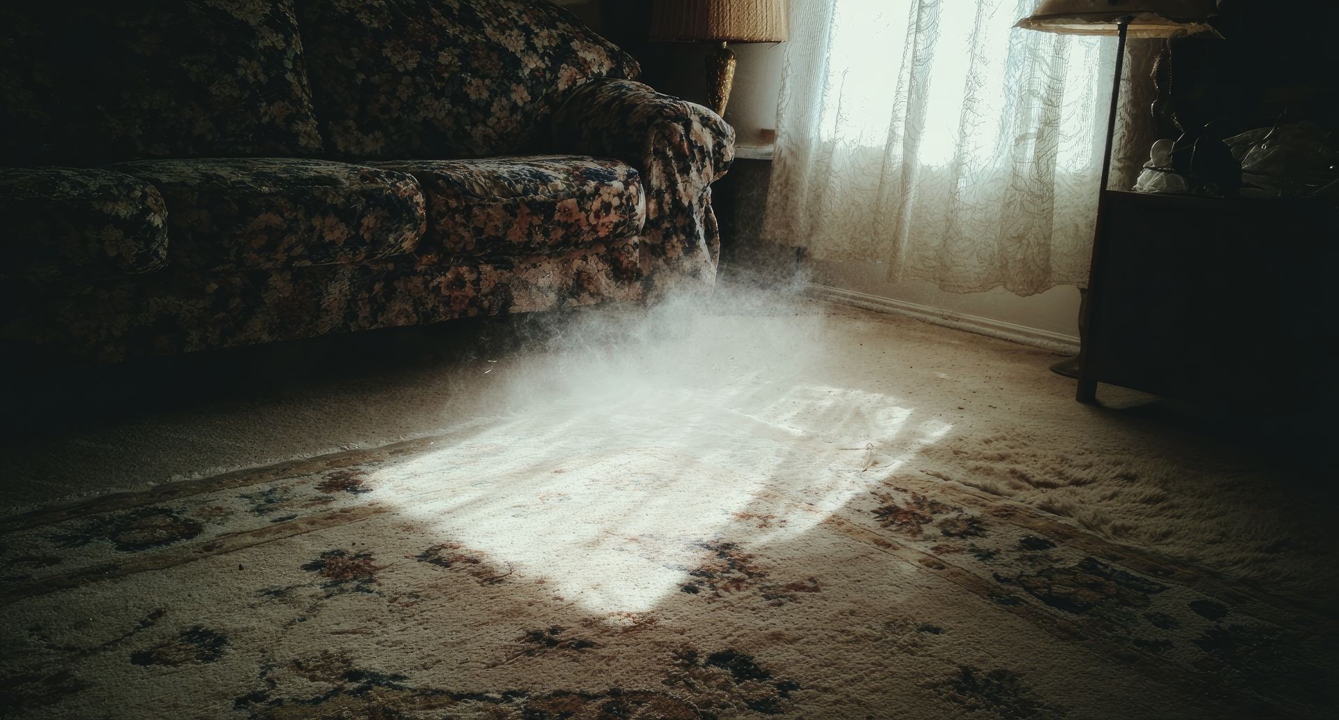 Des particules de poussière, illuminées par la lumière du soleil, ruissellent sur un tapis usé dans une pièce faiblement éclairée où se trouve un canapé.