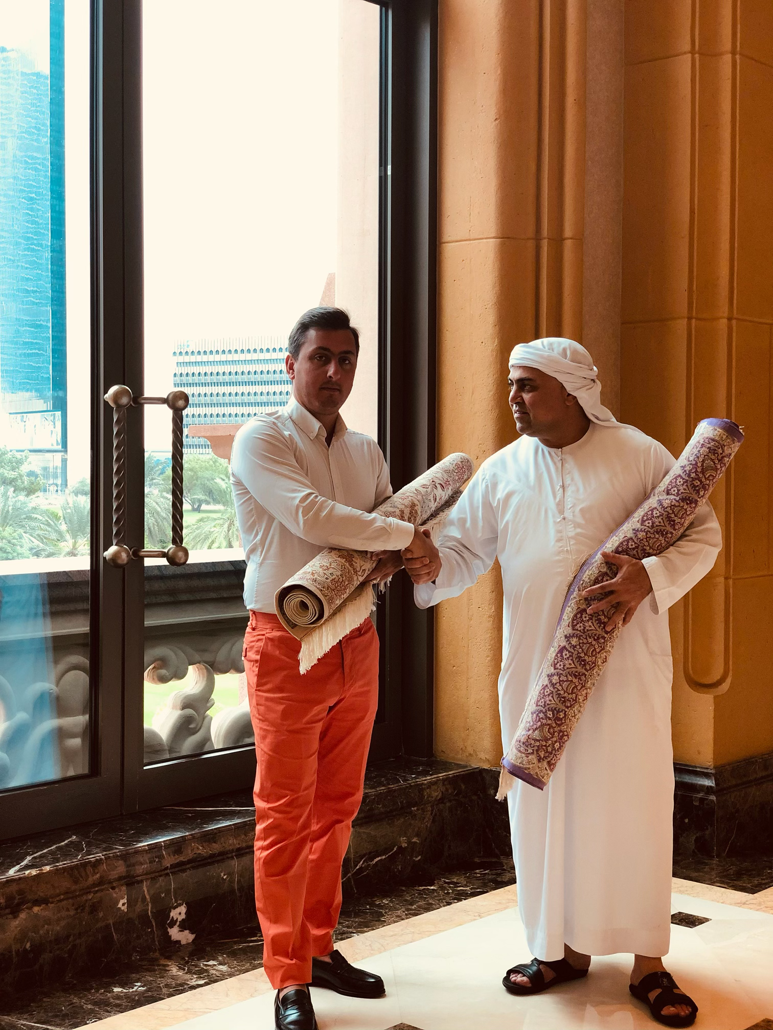 Cour intérieure avec colonnes arquées, grand tapis et plantes en pot. La lumière du soleil filtre à travers l'arche.