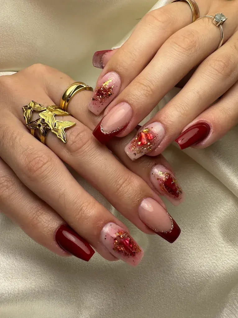 Manos con uñas cuadradas pintadas en rojo y estilo manicura francesa con detalles dorados y brillos.