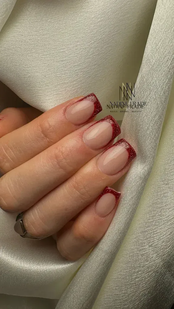 Mano con uñas cuadradas, manicura francesa con puntas de purpurina roja, apoyada sobre tela blanca.