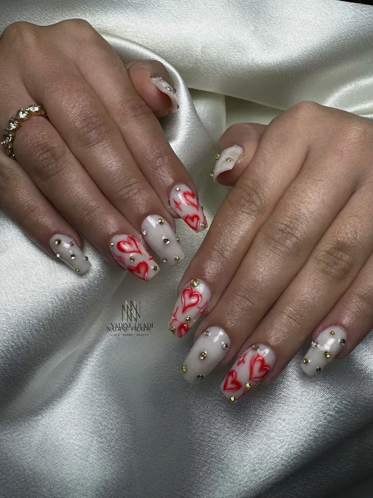 Manos con uñas claras, diseños de corazones rojos y tachuelas doradas, que descansan sobre satén blanco.