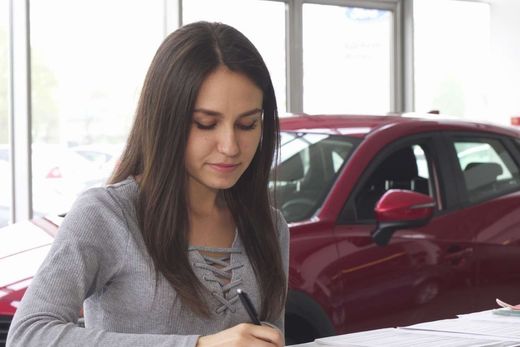 Una mujer está escribiendo en un trozo de papel delante de un coche rojo.