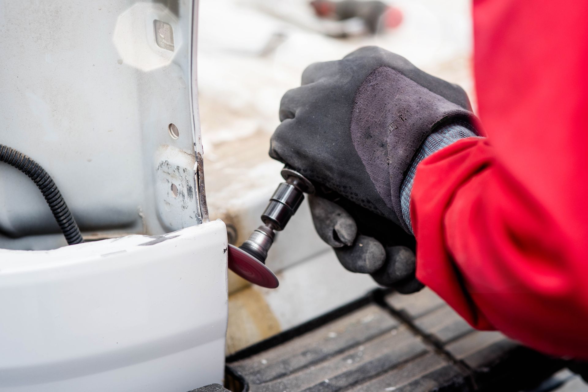 Une personne en veste rouge utilise un outil rotatif pour poncer une zone endommagée d'un véhicule blanc.