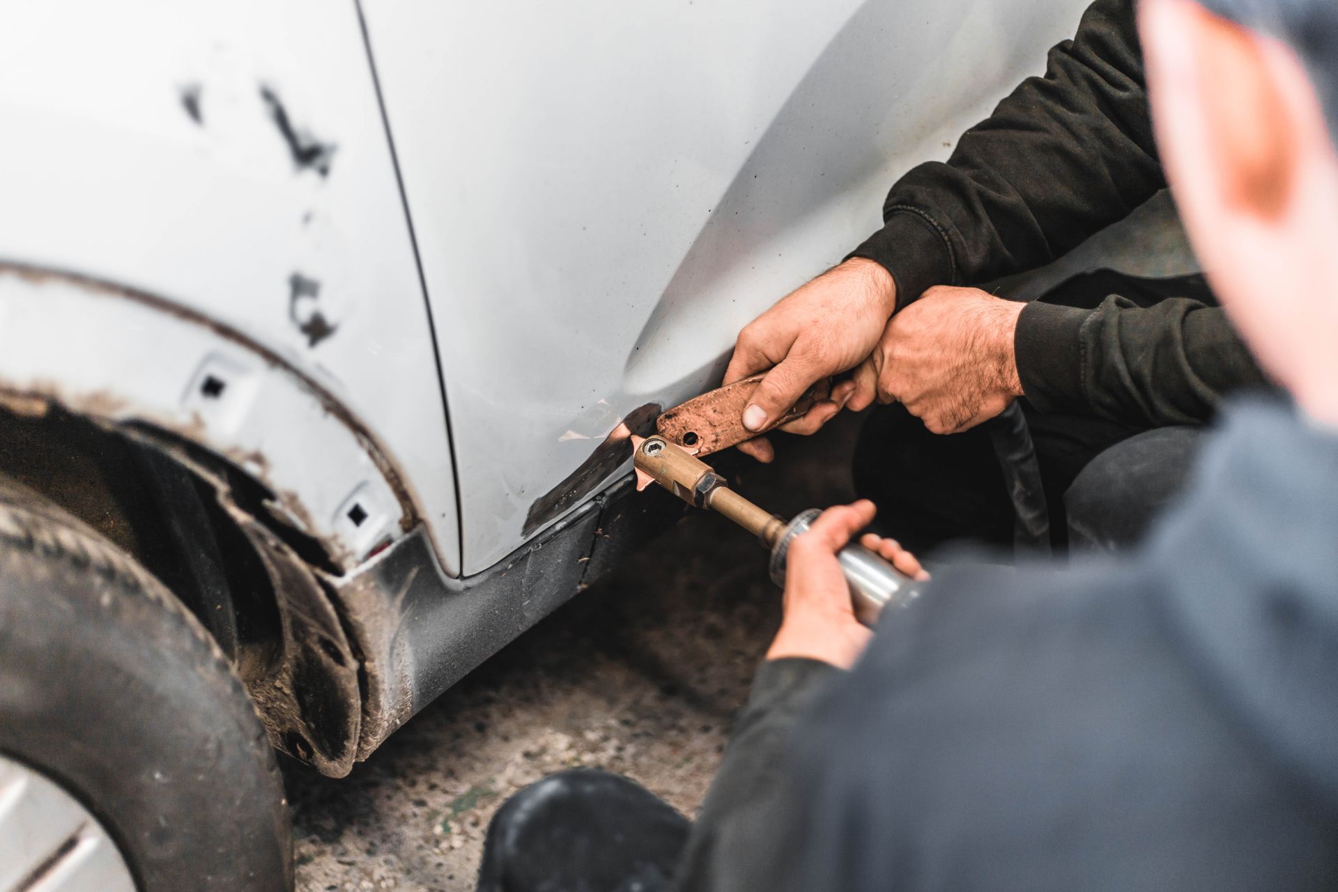 Deux mécaniciens réparent une voiture blanche ; l'un d'eux utilise un outil sur le panneau latéral.