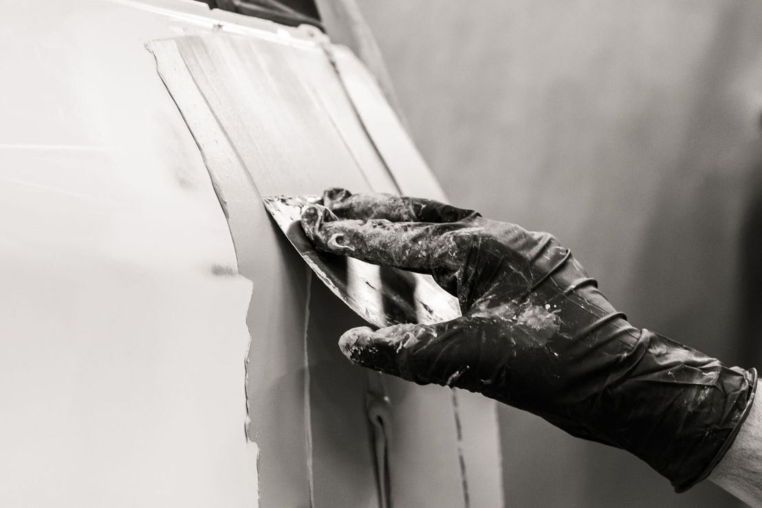 Main gantée lissant le mastic sur un panneau de voiture à l'aide d'un couteau à mastic.