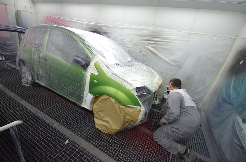 Une voiture est peinte en vert dans une cabine de peinture ; un ouvrier en costume gris applique la peinture.