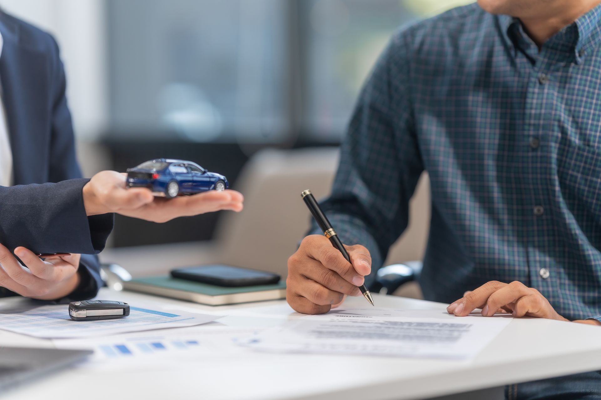 Une personne signe des documents à un bureau, une autre tient une petite voiture.