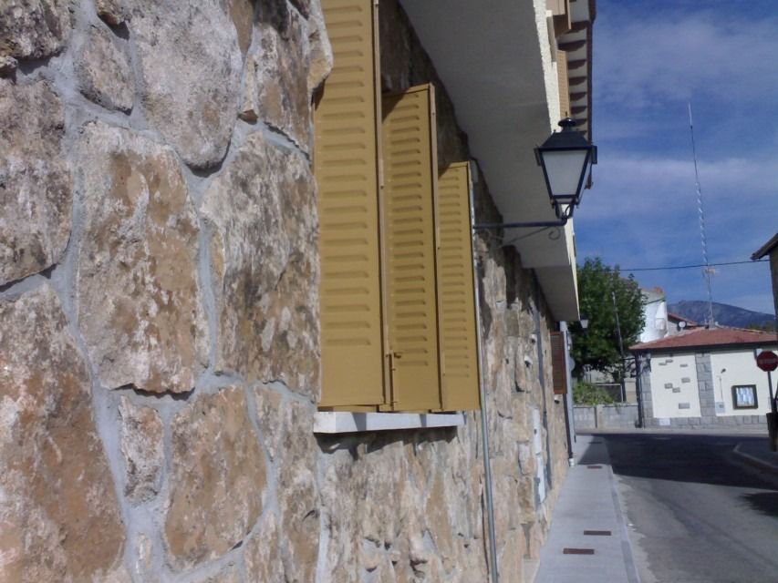 Muro de piedra con contraventanas amarillas a lo largo de una calle tranquila bajo un cielo azul.
