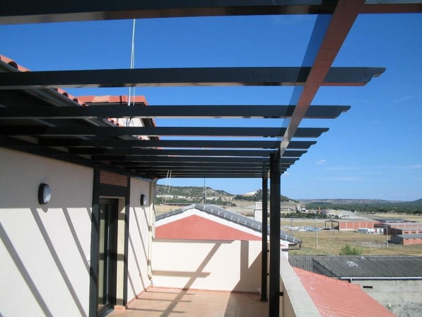 Balcón con vigas de pérgola oscuras con vistas a los tejados y a una ladera lejana bajo un cielo azul.