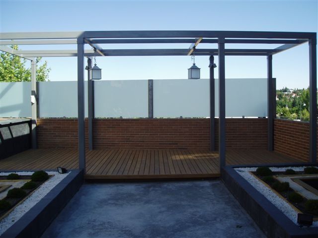 Patio moderno con terraza de madera, pérgola de metal oscuro y faroles colgantes, bordeado por paredes de ladrillo y color gris.