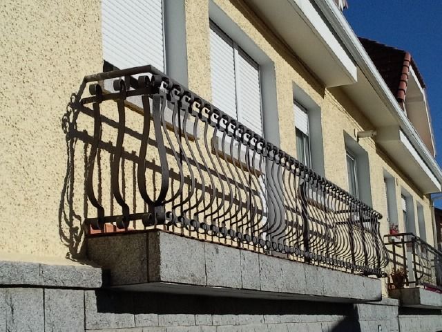 Barandilla de balcón de metal negro en el exterior de una casa con pared y ventanas de color beige.