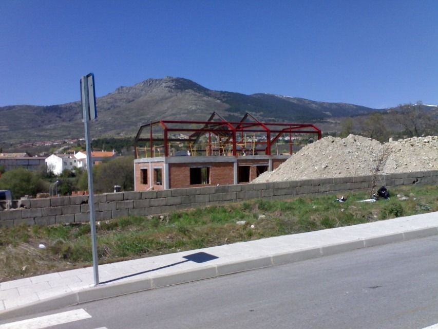 Estructura de ladrillo parcialmente construida con armazón de acero junto a una carretera, montañas y un cielo azul despejado al fondo.