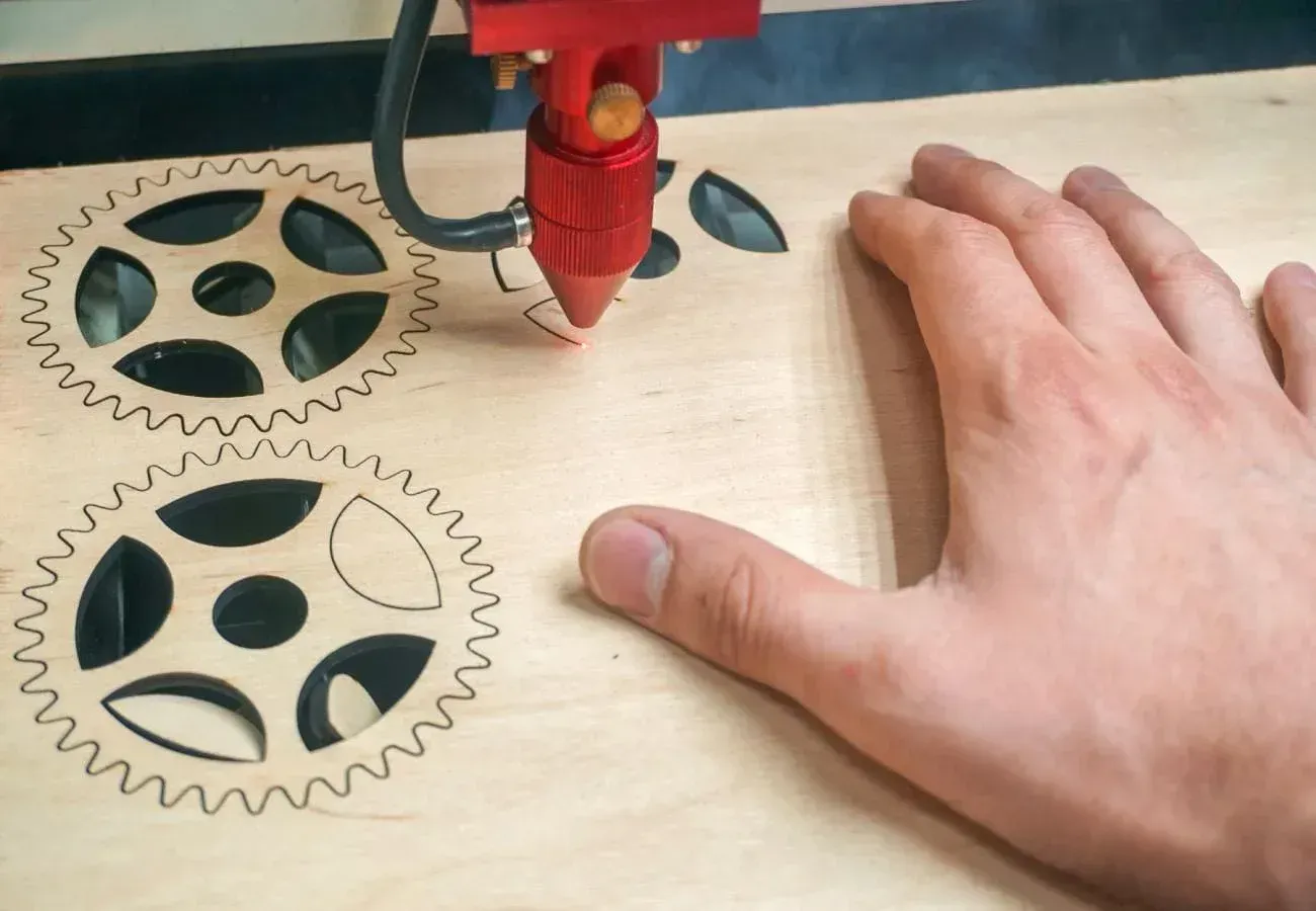 Una cortadora láser graba dos discos de madera con forma de engranaje mientras una mano sujeta el material.