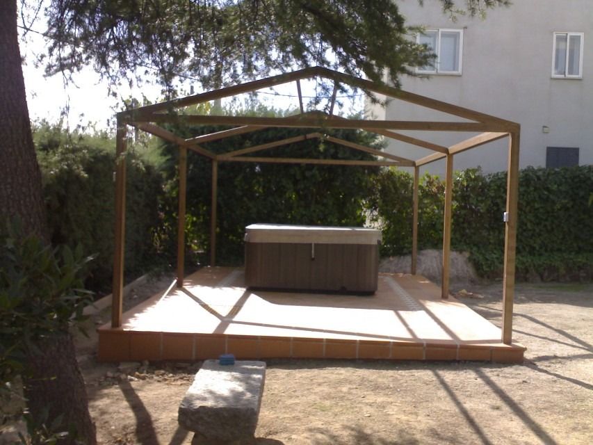 Estructura de madera tipo cenador en un patio soleado con una bañera de hidromasaje en el centro y árboles a su alrededor.