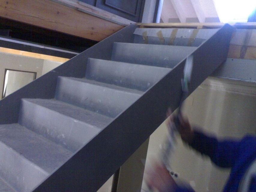 Escalera interior con escalones grises y el brazo de una persona sujetando la barandilla a la derecha.