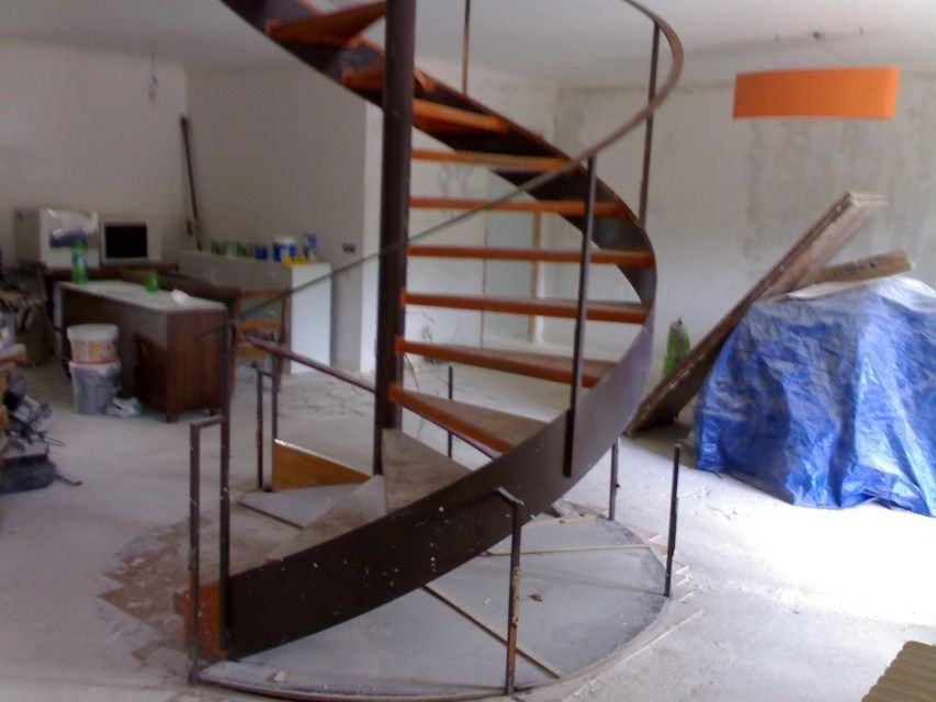 Escalera de caracol en construcción en una habitación sin terminar, con materiales cubiertos con lonas azules cerca.