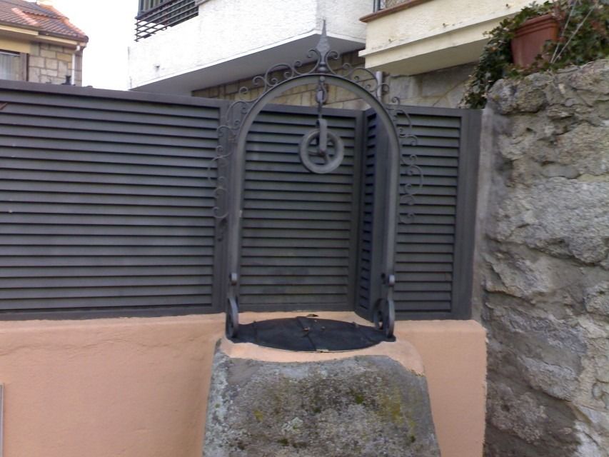 Pozo de piedra con asa de hierro frente a una puerta gris y un muro de piedra.