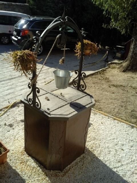 Pozo de los deseos decorativo con un cubo de metal y plantas secas sobre una base de madera para exteriores.
