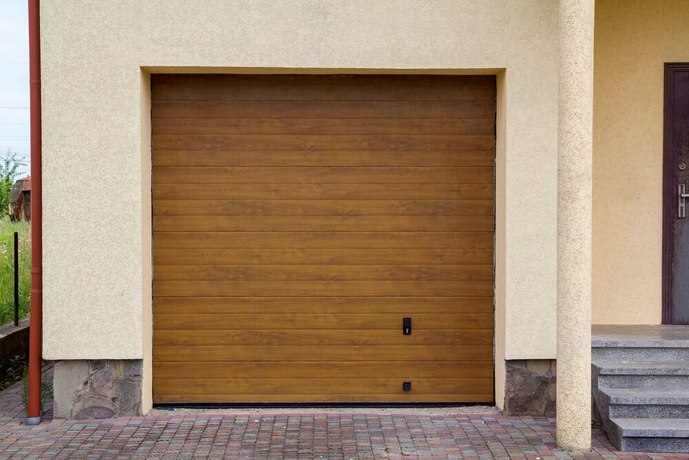 Puerta de garaje de madera cerrada en una pared de estuco beige junto a escalones y una entrada para vehículos.