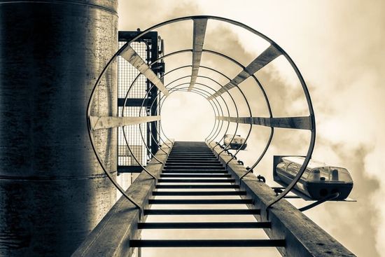 Una escalera metálica conduce a través de un túnel circular hacia una abertura iluminada, con un barco a la derecha.
