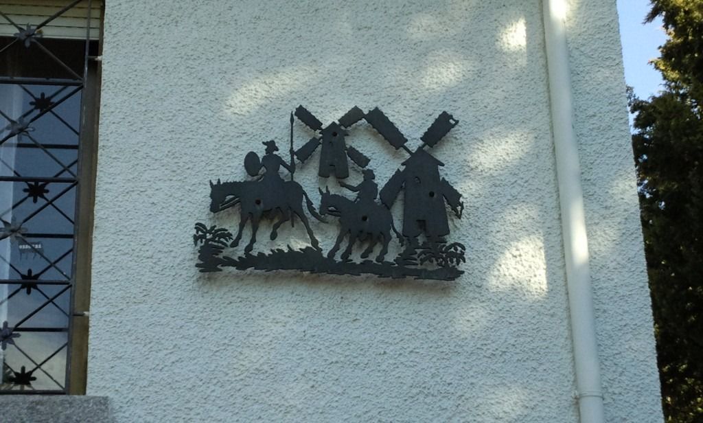 Escultura mural de metal que representa casas y una persona con un animal en la fachada de un edificio de estuco claro.