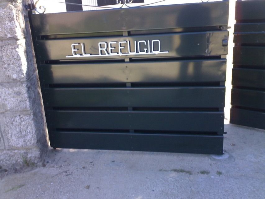 Puerta verde oscuro con el letrero “EL REFUGIO” junto a un muro de piedra y suelo de hormigón.