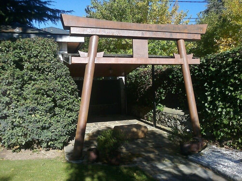 Puerta de jardín de madera con viga inclinada, enmarcada por arbustos y árboles bañada por la luz del sol.