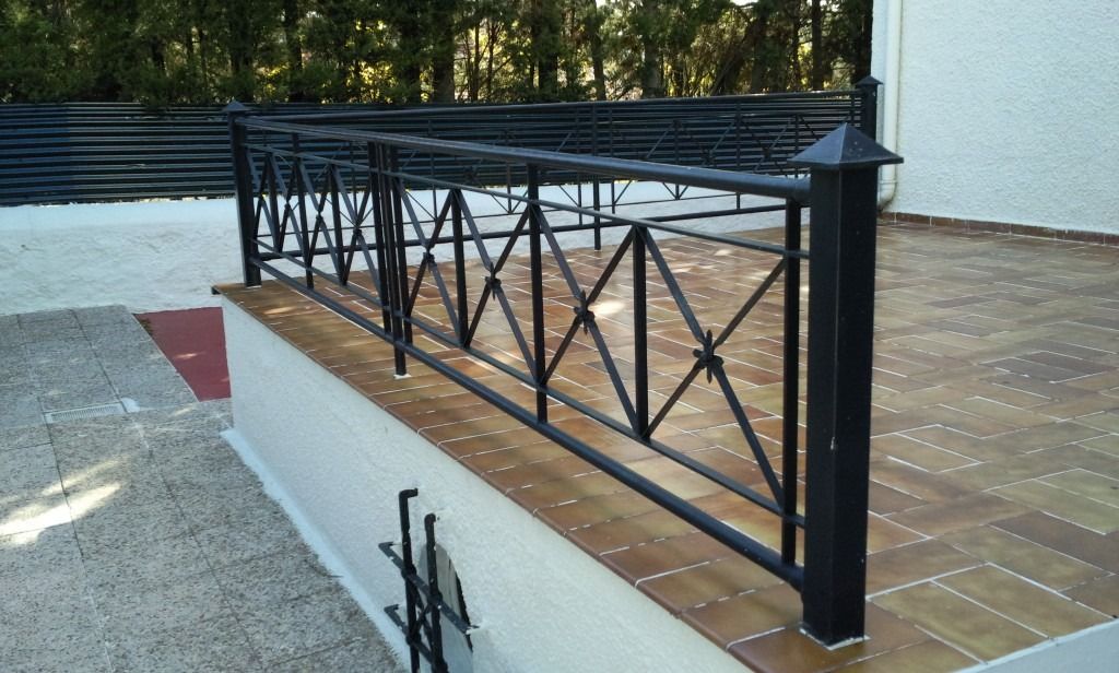Barandilla de metal negro a lo largo de una pasarela elevada de ladrillo junto a un camino pavimentado y árboles.