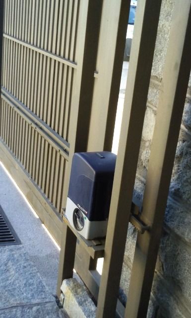 Primer plano de una puerta metálica de color beige con un pestillo automático azul y barrotes verticales cerca de una acera.