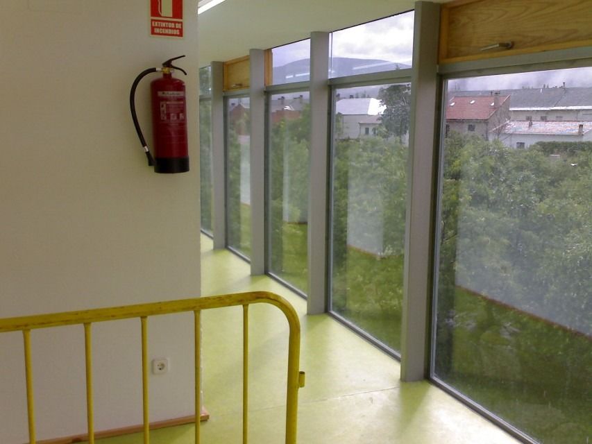 Pasillo luminoso con ventanales que van del suelo al techo, barandilla amarilla y un extintor rojo en la pared.