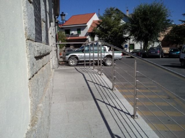 Acera junto a un muro de piedra con barandilla de metal, que conduce a un coche plateado aparcado en una calle soleada.