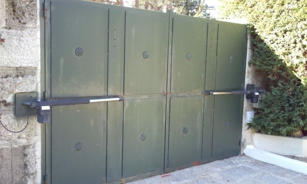 Gran puerta metálica verde en la entrada de un camino de acceso, con un pestillo negro y un muro de piedra a su lado.