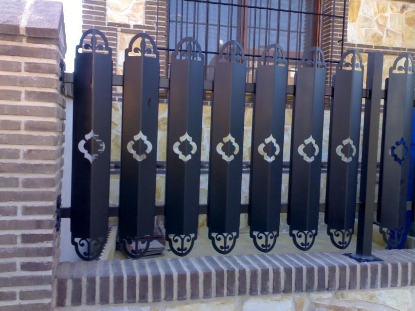 Valla decorativa de metal negro con detalles blancos frente a un muro de ladrillo y una puerta.