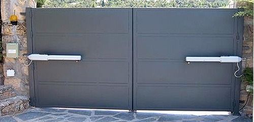 Puerta doble gris cerrada con dos manijas plateadas horizontales, ubicada entre muros de piedra y pavimento.