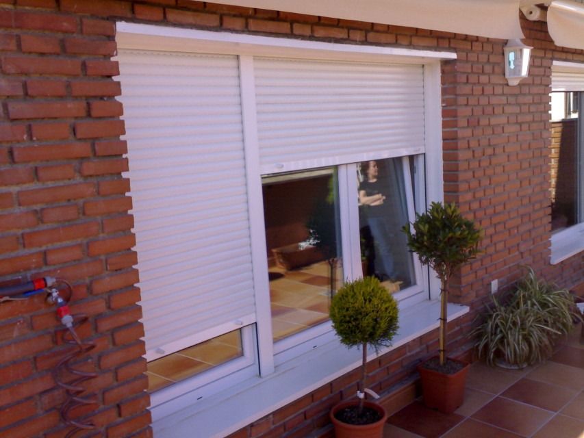 Ventana de una casa de ladrillo con contraventanas blancas y plantas en macetas en un patio.