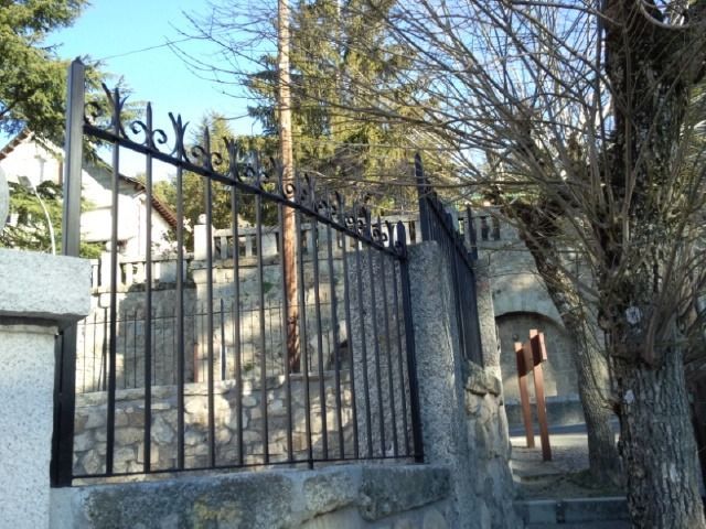 Una verja de hierro negro junto a escalones de piedra y un árbol en un sendero soleado en la ladera de una colina.