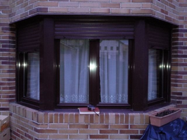 Ventanal de una casa de ladrillo con molduras de madera oscura y cortinas blancas; dos pequeños objetos rojos en el alféizar.
