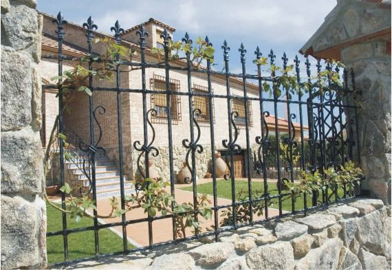 Casa de piedra vista a través de una elaborada verja de hierro forjado negro, con jardín y escaleras al frente.