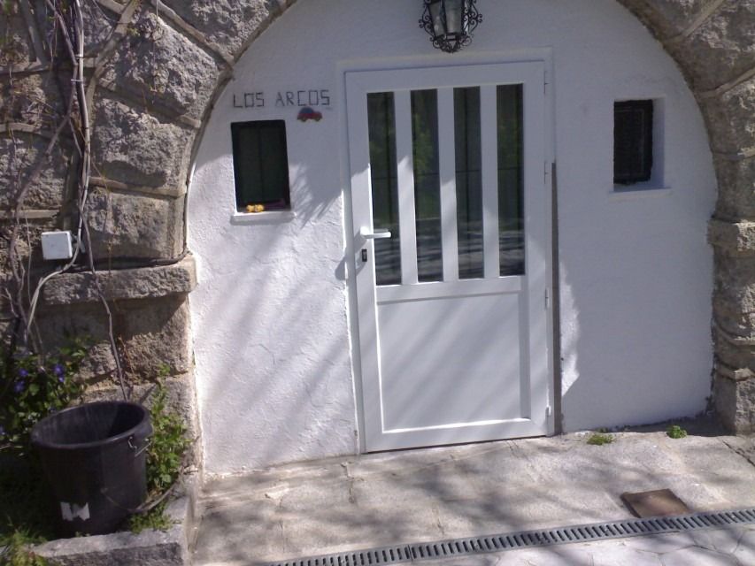 Puerta blanca enmarcada por un arco de piedra con una pequeña ventana y un cubo de basura negro al lado.
