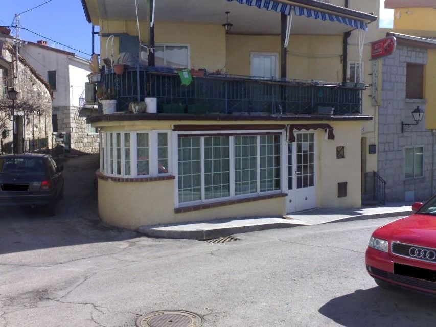 Vista de la calle de una casa amarilla de dos pisos con balcón, ventanales blancos y coches aparcados cerca.