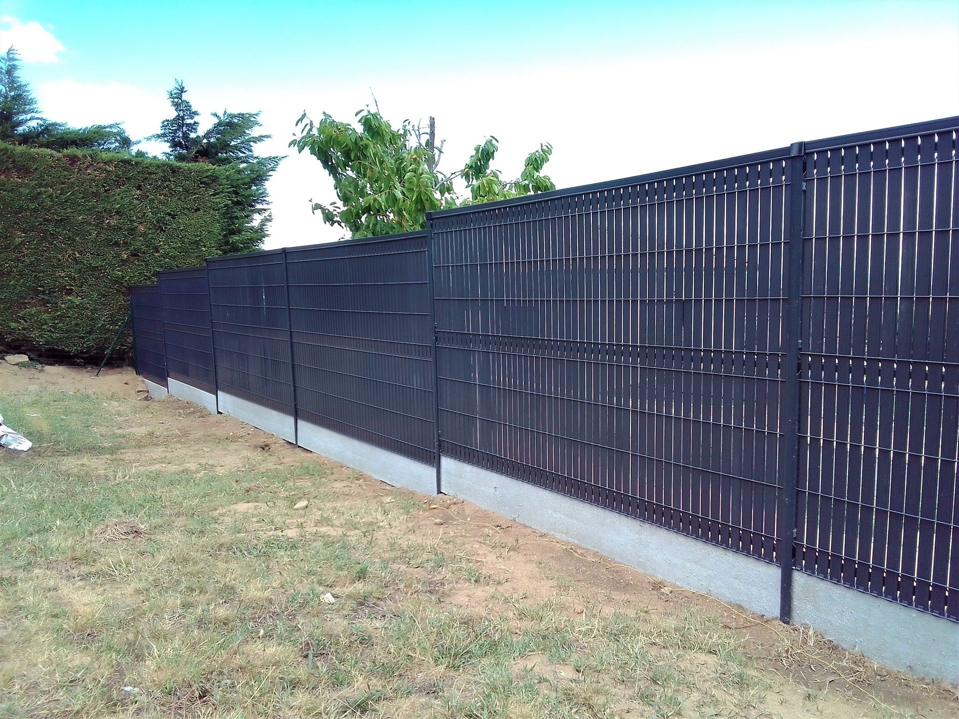 Clôture à lattes horizontales brunes avec des poteaux foncés, bordant un trottoir à côté de buissons et d'arbres verts.