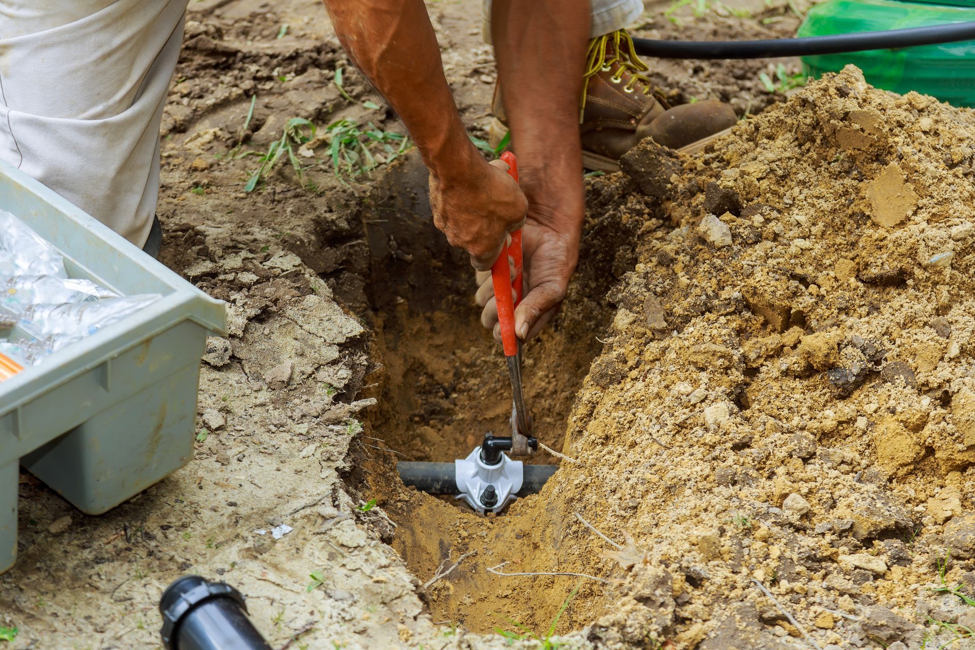 Une personne utilise une pince pour travailler sur la plomberie dans un trou creusé dans la terre à l'extérieur.