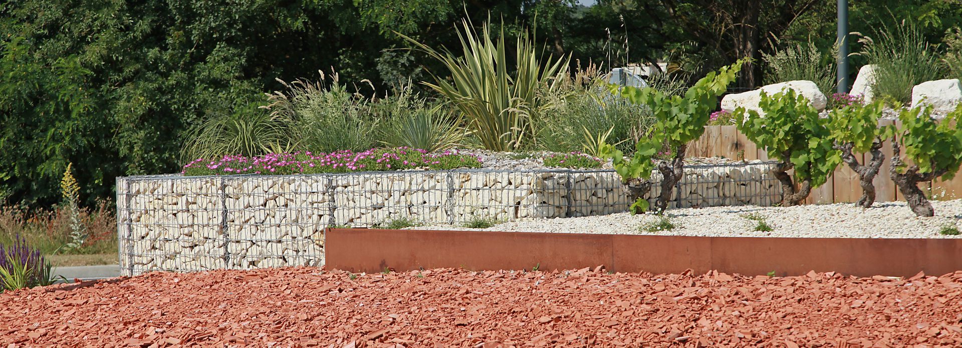 Jardin paysager avec mur en gabions, plantes et gravier rouge.