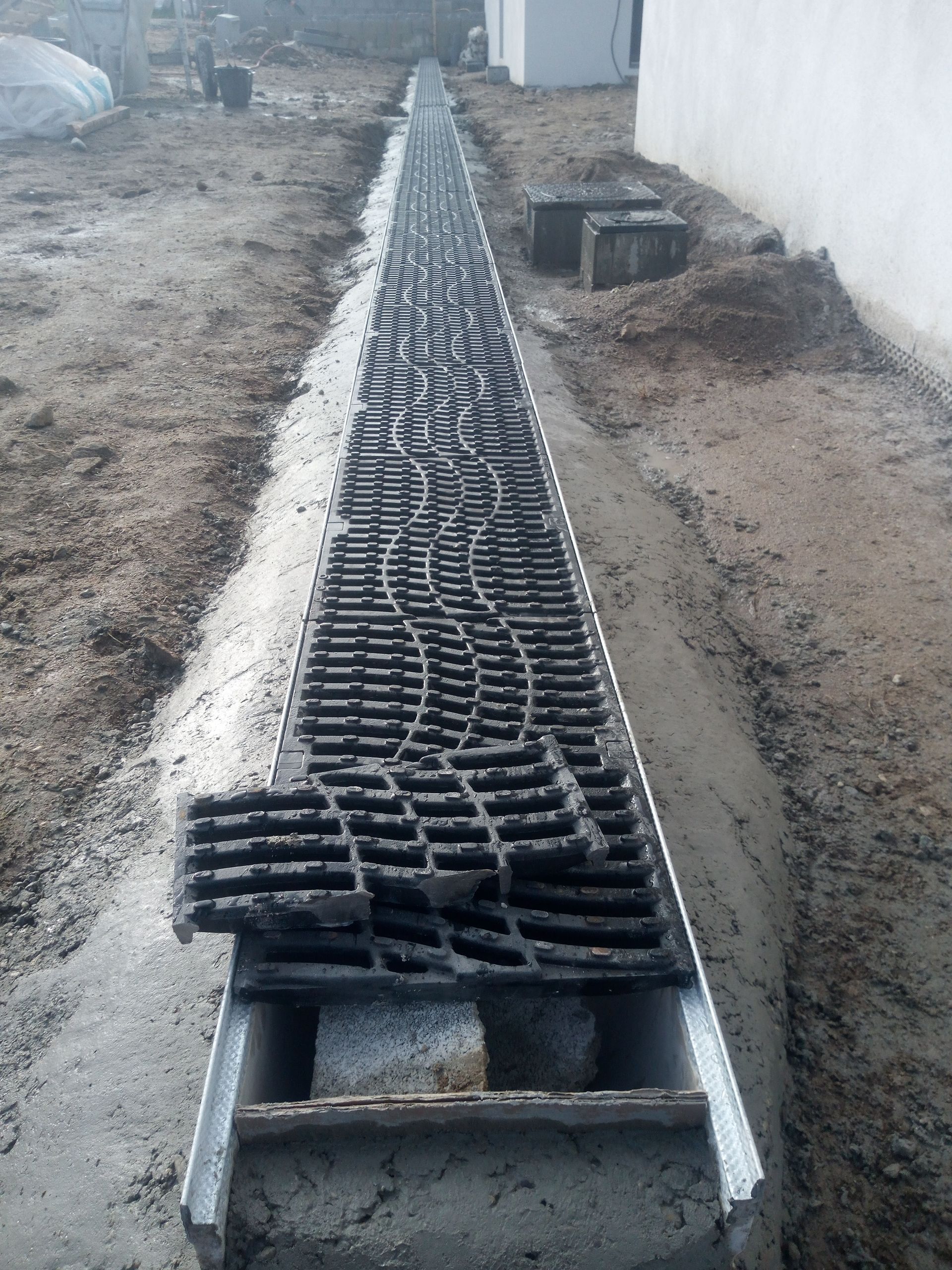 Un long caniveau de drainage muni d'une grille métallique, installé le long d'un bâtiment en construction.