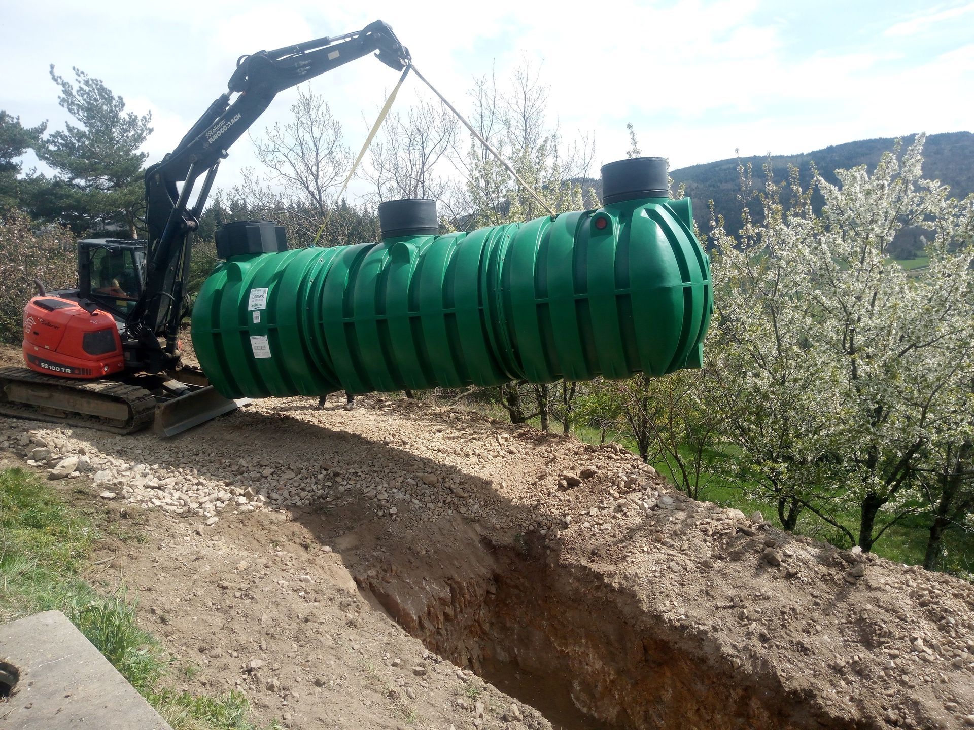 Une excavatrice descend une grande cuve verte dans une tranchée, probablement pour une fosse septique ou le stockage d'eau.