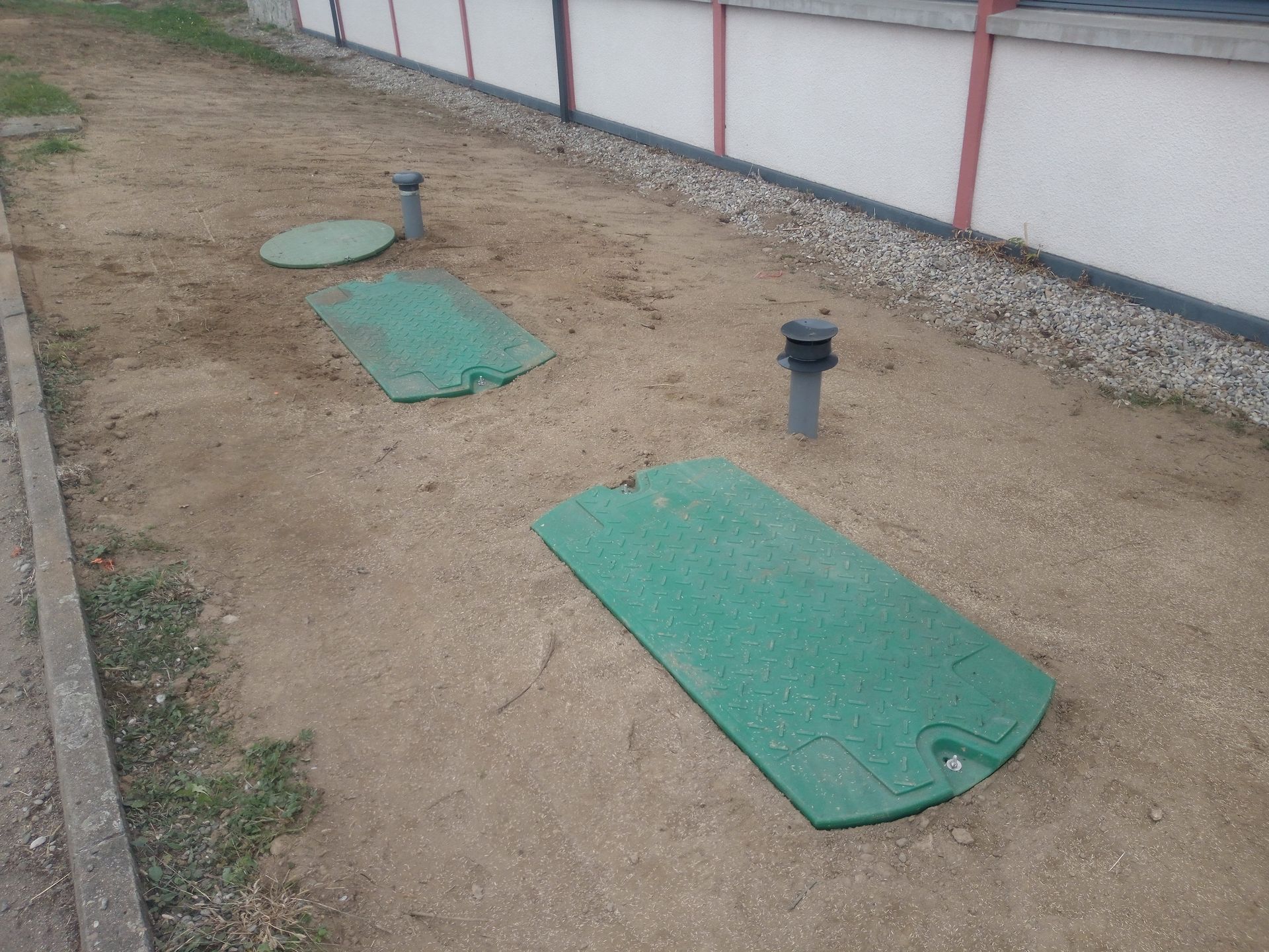 Trois plaques vertes et deux tuyaux gris sur un sol sablonneux à côté d'un bâtiment.